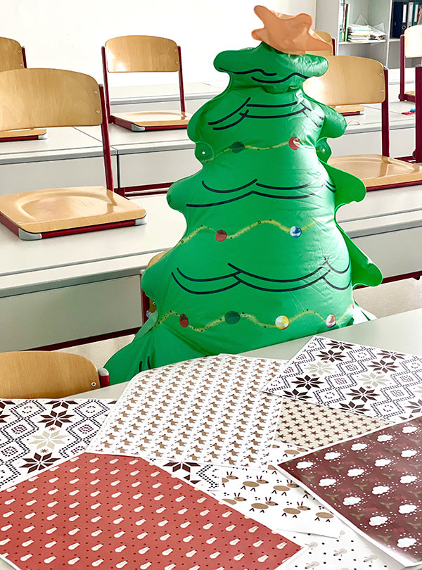Geschenkpapier liegt auf Schülertisch, dahinter aufblasbarer Christbaum