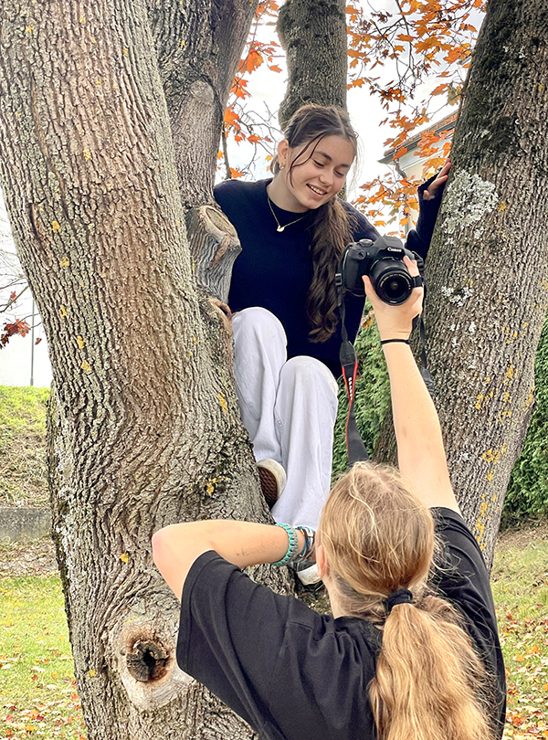 Schülerin im Baum sitzend schaut auf Bild in der Kamera, die ihr eine Schülerin entgegenstreckt