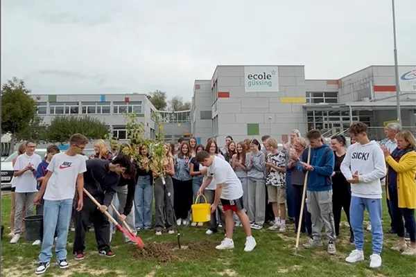 Das Schulgebäude der ecole güssing, davor die Schulgemeinschaft beim Pflanzen eines Baumes